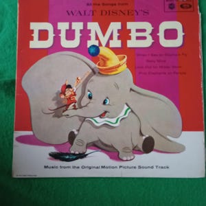 Pode incluir: Capa de álbum de vinil vintage de "Dumbo" da Walt Disney. A capa apresenta uma ilustração de desenho animado de Dumbo, o elefante, usando um chapéu amarelo com um pompom azul. O título "DUMBO" está em letras brancas grandes.