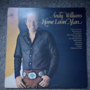 May include: Vintage vinyl record album cover featuring Andy Williams, titled "Home Lovin' Man." The cover shows Williams in a dark blue shirt and brown pants, with a decorative belt buckle. The album's song titles are listed on the right.