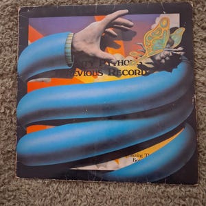 May include: A square vinyl record sleeve featuring a surreal illustration of a hand reaching towards a colourful butterfly. The text reads Monty Python's Previous Record. A large blue tube-like shape wraps around the centre against an orange background.