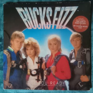 May include: Album cover for Bucks Fizz's "Are You Ready." The image shows four people in colorful outfits and parachutes. The album title is in large, stylized letters. The cover also includes the names of the hit singles.