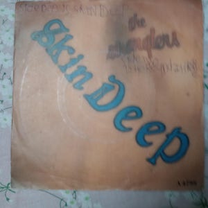 Puede incluir: Una funda de disco vintage con la inscripción "Skin Deep" en letras turquesas grandes. El fondo es de color melocotón. El texto adicional incluye "Side den is Skin Deep" y "the stranglers".