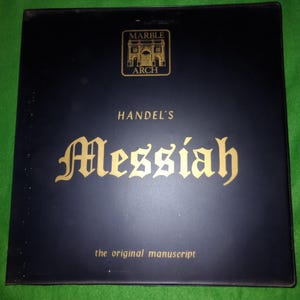 Op de afbeelding: Een zwarte map met gouden letters en een gouden embleem. De tekst op de map luidt "Marble Arch", "Handel's Messiah" en "the original manuscript". De map ligt op een groene ondergrond.