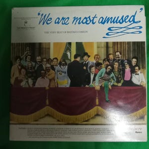 Può includere: Copertina di un album in vinile vintage intitolata "We are most amused", sottotitolata "The Very Best of British Comedy". La copertina mostra un gruppo di persone su un palco, tra cui una persona in costume da Batman.