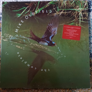 Puede incluir: Portada del álbum "The Complete Mike Oldfield" con un pájaro en vuelo sobre el agua. El nombre del artista y el título del álbum son visibles. Una pegatina roja enumera los títulos de las canciones, incluyendo "Moonlight Shadow".
