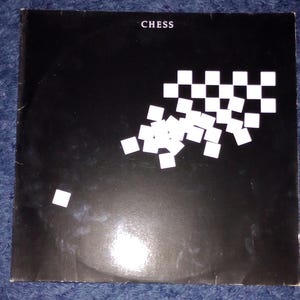 Könnte beinhalten: Ein schwarzes Albumcover mit dem Wort "CHESS" in Weiß oben. Das Cover zeigt eine verstreute Anordnung von weißen und schwarzen Quadraten, die einem Schachbrett ähneln.