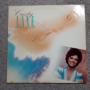 Könnte beinhalten: Albumcover für Cristy Lanes "Footprints in the Sand". Das Cover zeigt ein Porträt einer Frau und eine Sandstrandszene mit Fußabdrücken. Titel und Künstlername sind in eleganter Schrift gehalten.