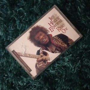 Può includere: Una musicassetta con l'album "Jimi Hendrix Experience Radio One". La copertina presenta una foto di Jimi Hendrix che suona la chitarra. La cassetta è su una superficie verde e strutturata.