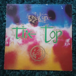 May include: Album cover for "The Top" by The Cure. The cover features a colorful abstract design with the band's name and album title in green and gold lettering. The background has blue, red, and yellow hues.