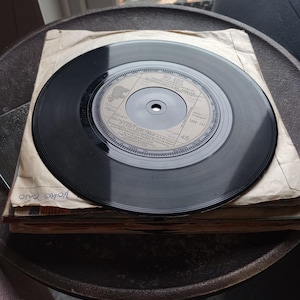 May include: A black vinyl record with a paper sleeve, resting on a stack of similar records. The record label is visible, with text including "BEE GEES" and "I Started A Joke". The records are on a dark, textured surface.