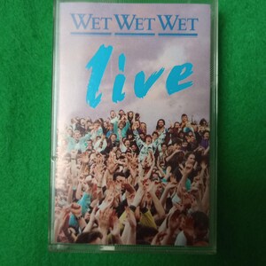 Puede incluir: Una cinta de cassette con la portada del álbum "Wet Wet Wet Live". La portada presenta el nombre de la banda en azul, con la palabra "live" en una fuente grande y estilizada. El fondo muestra una multitud de personas con las manos levantadas.