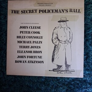 May include: Album cover for "The Secret Policeman's Ball" presented by Amnesty International. The cover has a black and white illustration of a man in a trench coat and hat. The cast includes John Cleese, Peter Cook, and Rowan Atkinson.