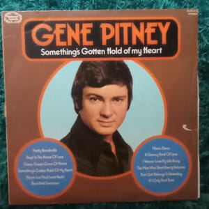 Op de afbeelding: Vintage albumhoes van Gene Pitney met de titel "Something's Gotten Hold of My Heart" in oranje en zwart. De hoes toont een portret van de zanger. Songtitels staan in blauwe cirkels.