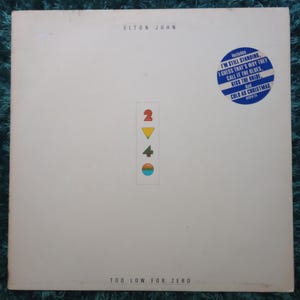 以下が含まれることがあります： エルトン・ジョンの「トゥー・ロウ・フォー・ゼロ」のアルバムカバー。カバーは主に白で、アーティスト名が上部にあります。曲名が書かれた青いステッカーと、カラフルな図形グラフィックが特徴です。アルバムタイトルは下部にあります。
