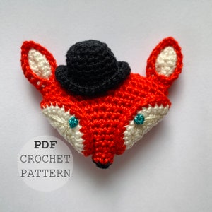May include: A crocheted fox wearing a black top hat with teal eyes. The fox is red and white with a black nose. The text "PDF CROCHET PATTERN" is in a circle below the fox.