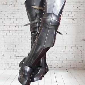 Puede incluir: Armadura de pierna de cuero negro, de altura de rodilla, con detalles metálicos y correas. La armadura se lleva sobre unos vaqueros y una camisa de cuadros.