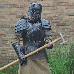Puede incluir: Una armadura medieval negra completa, que incluye un casco, peto, protectores de hombros y protección para las piernas. La armadura se usa con una camisa de cota de malla y una falda color canela. Se sostiene un martillo de mango largo.