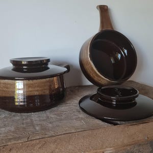 Puede incluir: Un juego de utensilios de cocina de cerámica marrón vintage, que incluye una olla pequeña con tapa, una cacerola con asa y una tapa. Los utensilios de cocina tienen un acabado brillante y una combinación de colores marrón de dos tonos. Los artículos se exhiben sobre una superficie de madera.