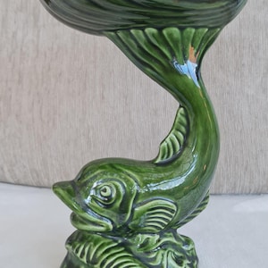 May include: A green ceramic dolphin-shaped decorative bowl. The bowl has a wide, scalloped top and a curved stem that forms the dolphin's body. The dolphin rests on a textured base, and the entire piece has a glossy finish.