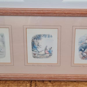 May include: A framed artwork featuring three framed illustrations. The illustrations depict scenes with figures in a soft color palette. The frame is a light brown color. The artwork is displayed on a wooden surface.