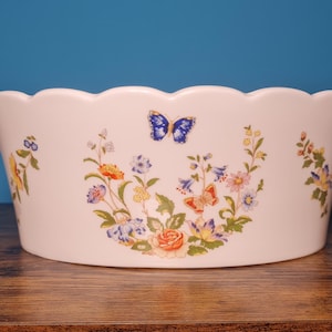 May include: White ceramic planter with a scalloped rim and handles. The planter is decorated with a butterfly and floral design in shades of blue, orange, yellow, and purple. The planter is on a wooden surface.