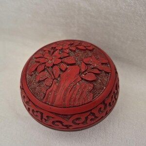 May include: A round red cinnabar-style lacquer box featuring a carved relief of flowers and rock formations on the lid. The sides of the box are decorated with an intricate openwork scroll pattern, showcasing detailed craftsmanship and texture.