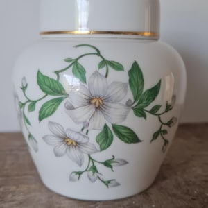 May include: White ceramic jar with a lid, decorated with a floral pattern of white flowers and green leaves. The jar has a gold band around the lid. The design is delicate and elegant, suitable for home decor.