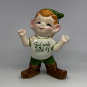 May include: A ceramic figurine of a smiling elf with orange hair and a green hat. The elf wears a white t-shirt with green trim and the text God made Irish #1 printed on the front, green shorts, and brown boots, with arms raised in a cheerful pose.