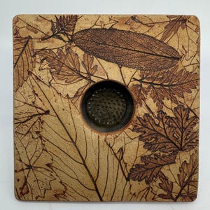 May include: Square ceramic vase with a brown and beige glaze. The vase is decorated with a pattern of pressed leaves in a dark brown color. The vase has a round opening in the center.