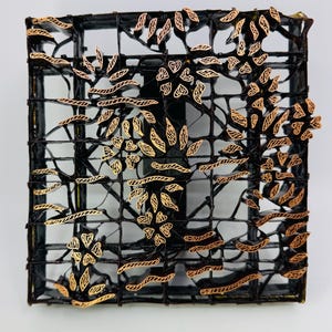 May include: A square batik printing stamp made of dark metal with a grid structure. The surface features intricate copper-colored floral and leaf patterns soldered onto the frame, designed for applying wax designs to fabric in textile printing.