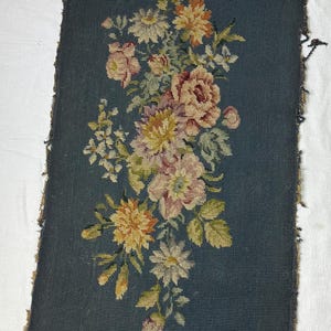 May include: A rectangular piece of needlepoint fabric with a dark teal background. The fabric features a floral design in shades of pink, yellow, and white. The edges of the fabric are frayed, showing signs of age.