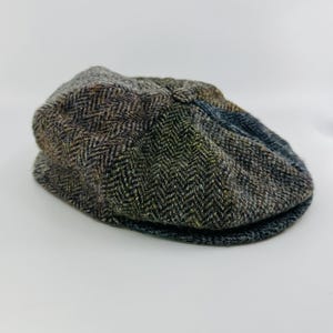 May include: A flat cap featuring a multi-colored herringbone pattern in shades of brown, olive green, and charcoal gray. The cap has a rounded crown and a short, stiff brim, constructed from a textured, woven fabric with a classic panel design.