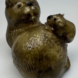 May include: A detailed figurine of a cat with a small mouse perched on its back. The cat is a muted olive green color with darker accents, and the mouse is a similar shade. The sculpture has a glossy finish and is likely made of ceramic or a similar material.