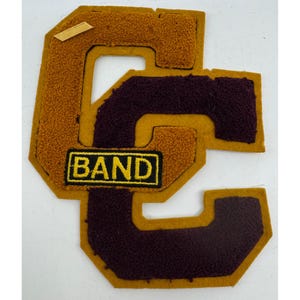 May include: A felt patch with a large gold "C" with a smaller maroon "C" inside. The patch has a small rectangular tag with the word "BAND" in yellow letters on a black background.