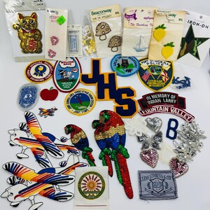 May include: A collection of assorted embroidered and sequined fabric patches featuring various designs including a gold cat, airplanes, a parrot, a star, lemons, mushrooms, a sailboat, and several circular emblems with text such as Port St. Lucie Police.