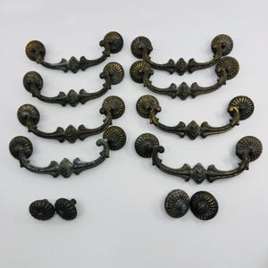 May include: A collection of six ornate metal drawer pulls with curved handles and circular mounting plates, featuring a dark bronze finish with gold-toned highlights, accompanied by four matching circular cabinet knobs with ribbed detailing.