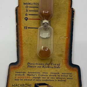 May include: Vintage Mueller's Macaroni Timer with a clear glass hourglass design. The timer has a brown sand and measures cooking times from 0 to 12 minutes. The timer is on a cardboard backing with text about macaroni products.