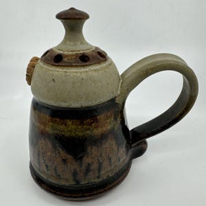 May include: A brown and white ceramic teapot with a cork stopper and a handle. The teapot has a brown and white glaze with a pattern of brown lines. The teapot has a lid with holes in it.