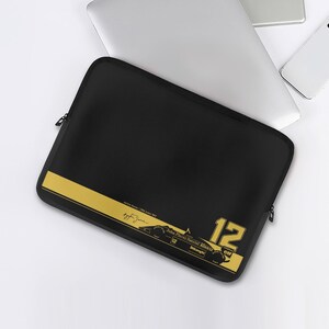 May include: Black laptop sleeve with a gold stripe and racing-themed graphics. The sleeve features the number 12 and text including "John Player Special Renault" and "Delonghi". A laptop, phone, and tablet are visible in the background.