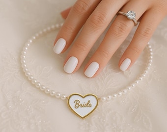 May include: A hand with white nails and a diamond ring, next to a pearl necklace with a heart-shaped pendant. The pendant has a gold border and the word "Bride" in gold script.