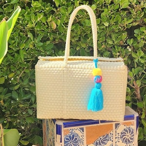 May include: A rectangular cream-colored woven tote bag with two matching handles. A decorative tassel featuring a bright yellow pom-pom, a pink bead, and a long light blue fringe is attached to one of the handles of the woven bag.