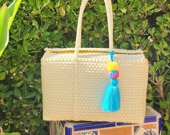 May include: A rectangular cream-colored woven tote bag with two matching handles. A decorative tassel featuring a bright yellow pom-pom, a pink bead, and a long light blue fringe is attached to one of the handles of the woven bag.