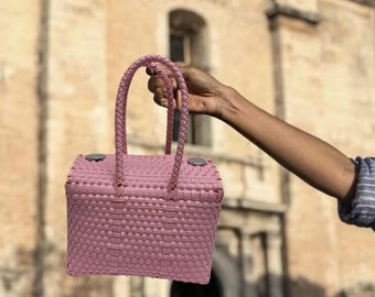 Puede incluir: Un bolso tipo cesta rectangular tejido en color rosa con forma estructurada y dos asas trenzadas. La parte superior del bolso presenta dos detalles metálicos circulares en tono plateado en las esquinas, que sirven como cierre para la tapa.
