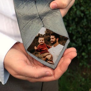 Puede incluir: Una corbata gris con una apariencia texturizada, que revela una fotografía de un hombre sosteniendo a un bebé. El bebé lleva un suéter rojo. La corbata se sostiene en las manos de una persona, con una camisa blanca y un gemelo visible.