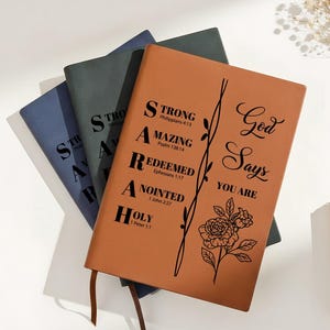 Puede incluir: Un conjunto de diarios encuadernados en cuero en tonos azul, verde y marrón. El diario marrón presenta el texto "God Says You Are" con ilustraciones florales y de vid. La palabra "SARAH" está impresa verticalmente en la portada, con palabras inspiradoras.