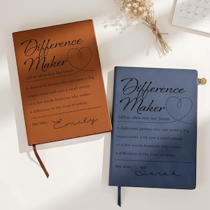 May include: Two leather-bound journals, one brown and one blue, are displayed. Each journal features the words "Difference Maker" and a definition, along with a name. A calendar and dried flowers are in the background.