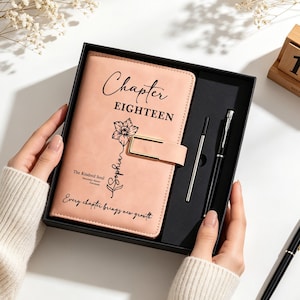 May include: A peach-colored journal in a black gift box, featuring the text Chapter EIGHTEEN, a floral illustration, the name Sophia, and the phrase Every chapter brings new growth. The set includes a black pen and a metal refill cartridge.