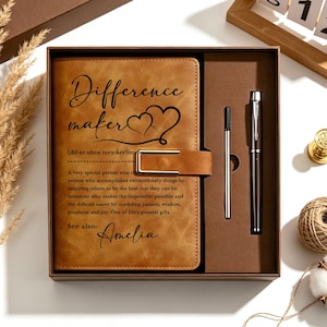 May include: A tan leather-look journal in a gift box with a matching pen and refill. The cover features the text "Difference maker" with a heart icon and a definition of the term, followed by the name "Amelia" in a cursive script font.