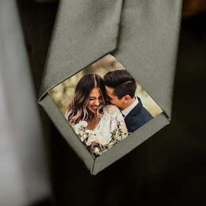 May include: A sage green tie featuring a personalized photo of a couple. The photo is a close-up of the couple, with the woman smiling and the man embracing her. The tie is held by a person, with a white shirt and dark suit visible.
