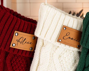 Op de afbeelding: Drie gepersonaliseerde kerstsokken in rood, crème en groen, elk met een kabelgebreid ontwerp. De sokken hebben een leren label met een naam: Alice, Eunice en Gladys. Ze hangen aan een witte schoorsteenmantel.