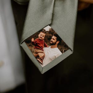 Może przedstawiać: Szałwiowa zielona krawat z personalizowaną wkładką ze zdjęciem przedstawiającą mężczyznę trzymającego dziecko. Krawat jest trzymany, prezentując niestandardowy obraz. Krawat jest wykonany z gładkiej tkaniny.